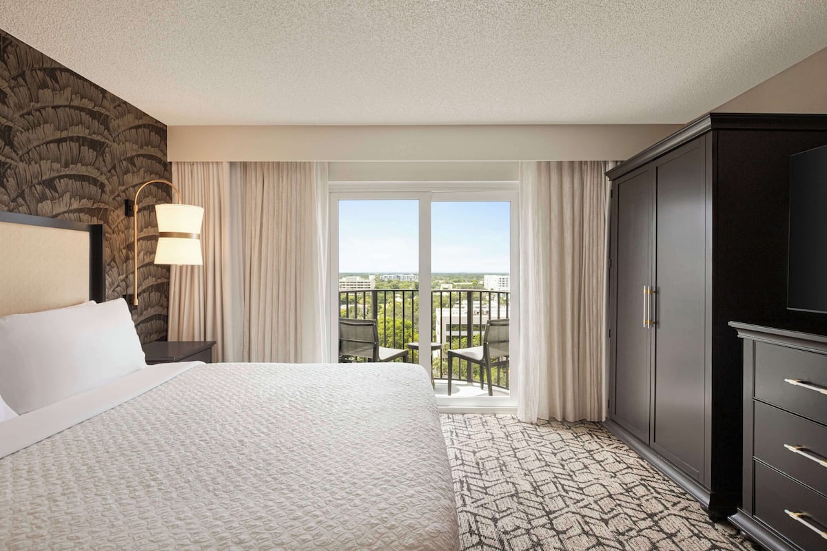 Ein Hotelzimmer mit einem großen Bett, einem Kleiderschrank, einem Nachttisch, einer Lampe und einem Balkon mit Blick auf die Stadt.