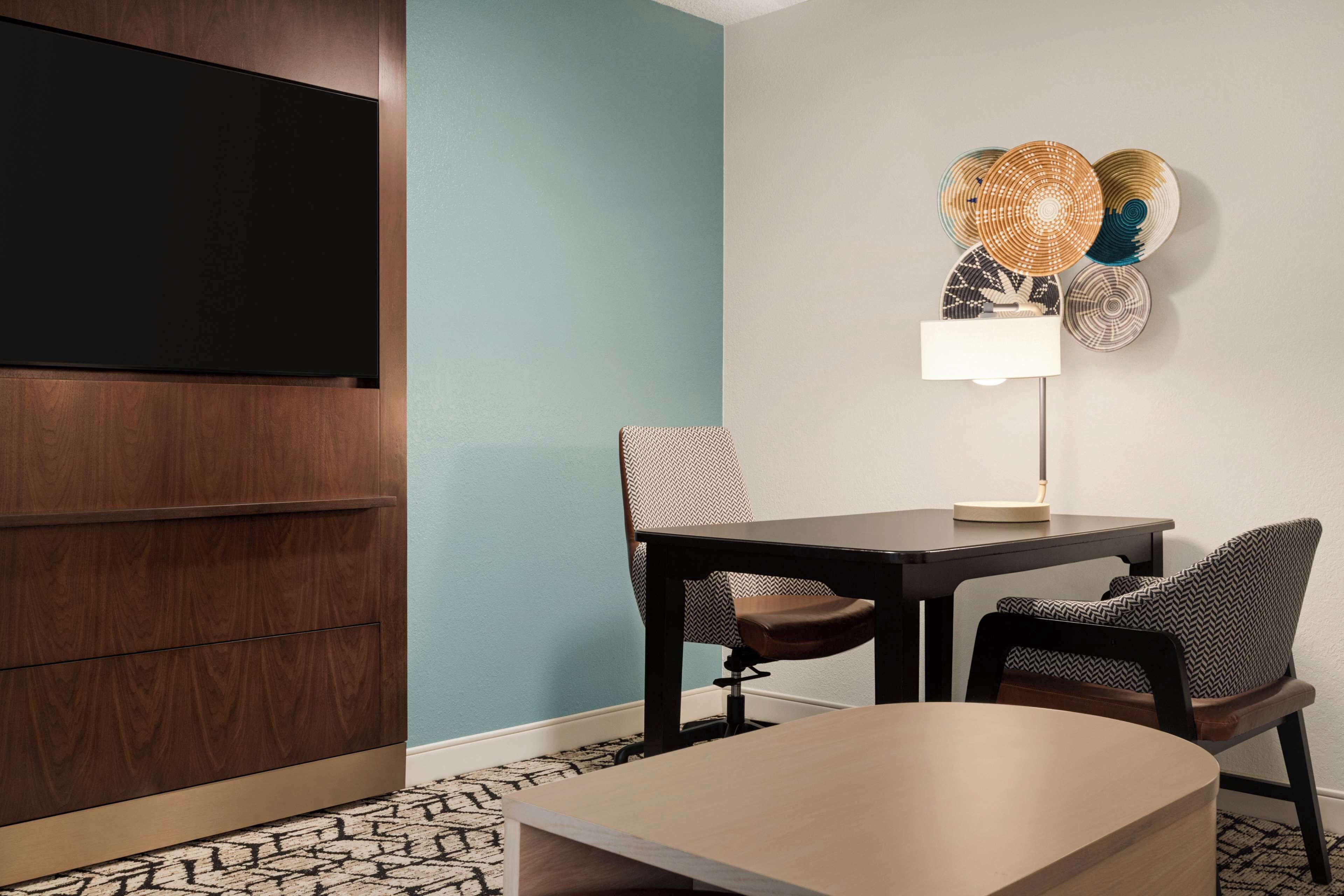 A hotel room with a flat-screen TV mounted on the wall, a dining table with two chairs, a lamp, and a decorative wall piece.
