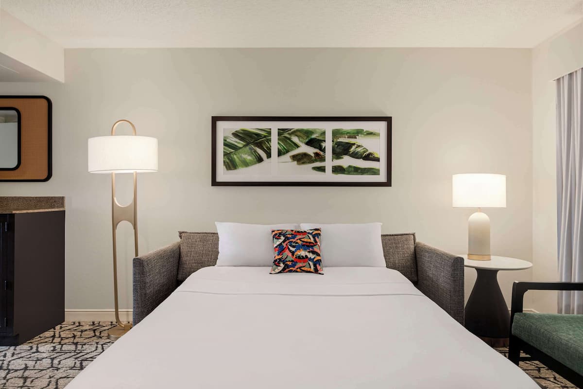 Ein Hotelzimmer mit einem großen Bett, zwei Nachttischlampen, einem gerahmten Kunstwerk mit einem tropischen Blatt, einem bunten Kissen, einem kleinen runden Tisch und einem grünen Stuhl.