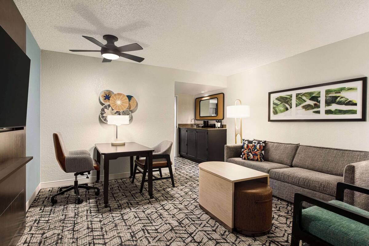 Ein Hotelzimmer mit Essbereich, einer Couch, einem Couchtisch, einem Deckenventilator, einer Küchenzeile und einem gerahmten Bild an der Wand.