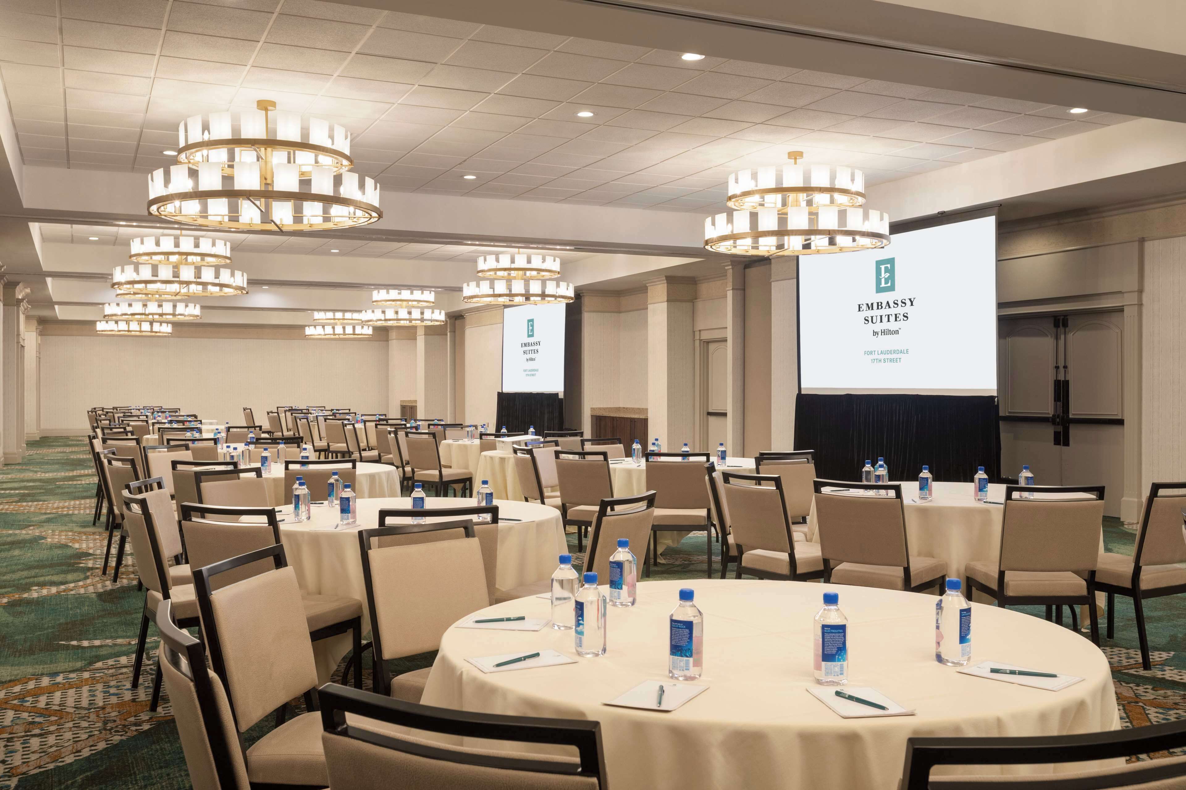 Un salón de conferencias con mesas redondas, sillas y botellas de agua, con una pantalla grande mostrando la marca 'Embassy Suites by Hilton'.