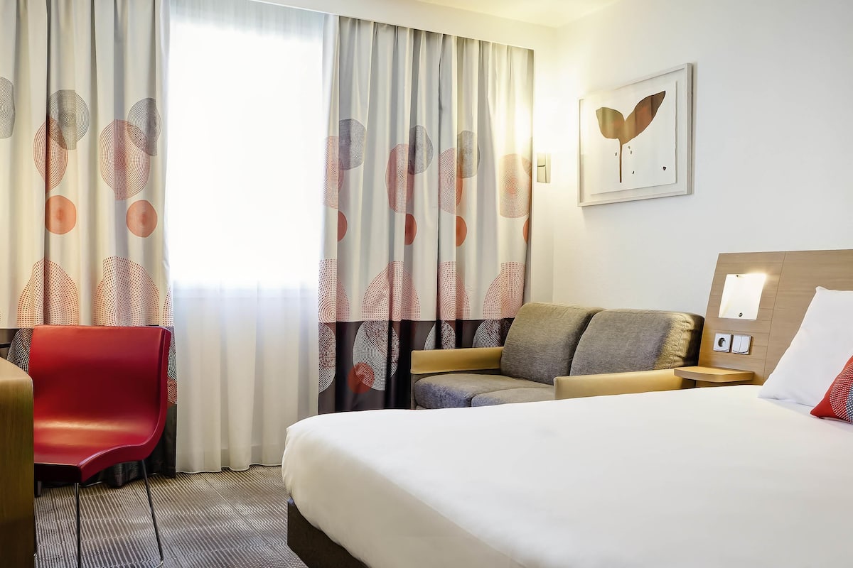 A hotel room with a bed, a sofa, a red chair, and a desk.