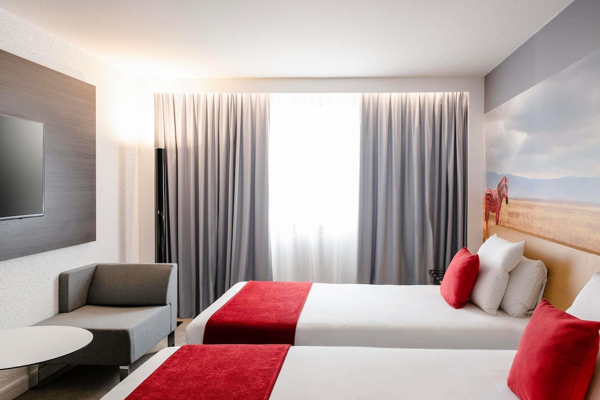 Hotellihuone, jossa on kaksi sänkyä, televisio, työpöytä ja suuri ikkuna, jossa on verhot.