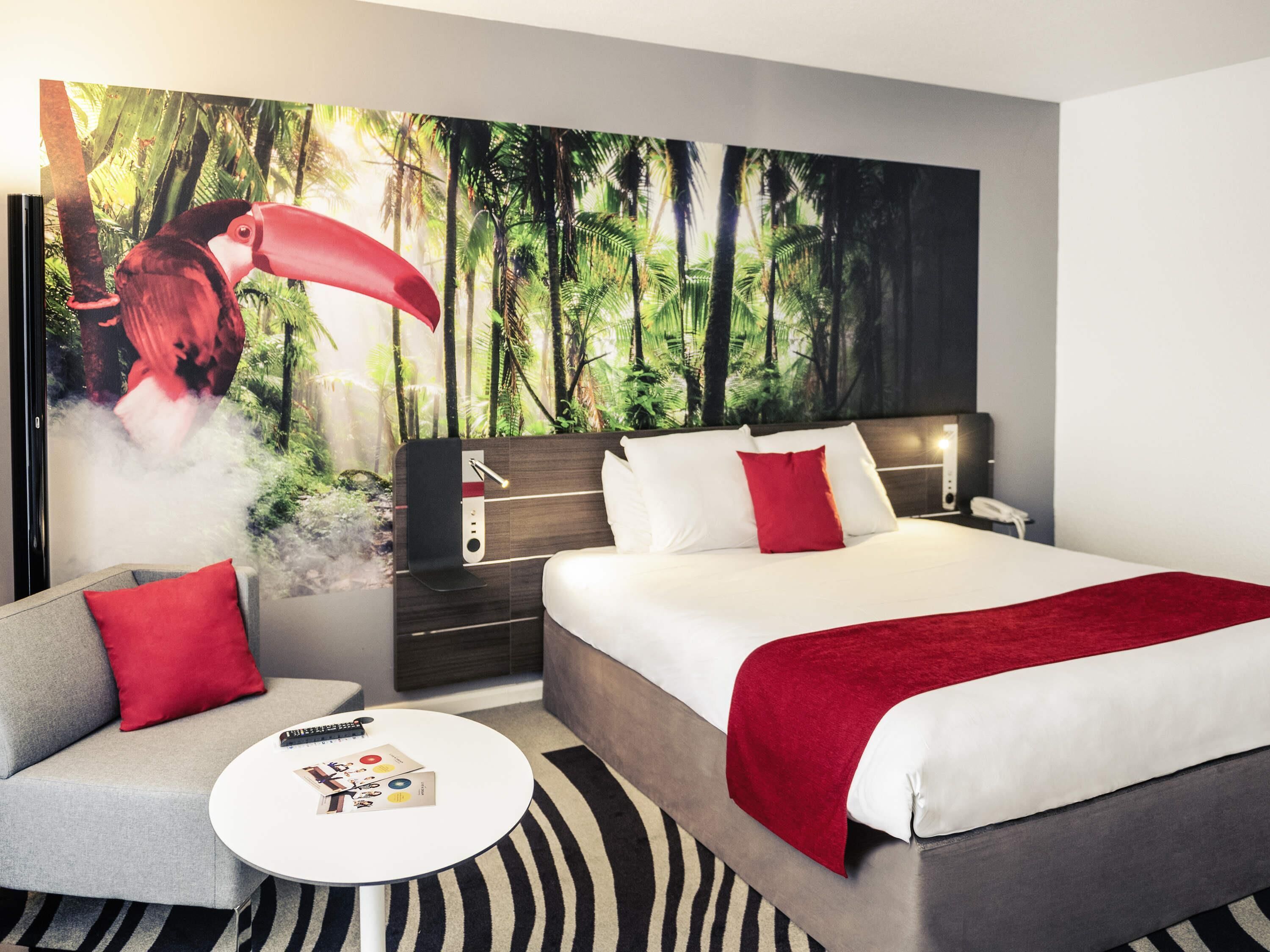 A modern hotel room with a large bed, a sofa, a small round table, and a wall mural of a forest scene.