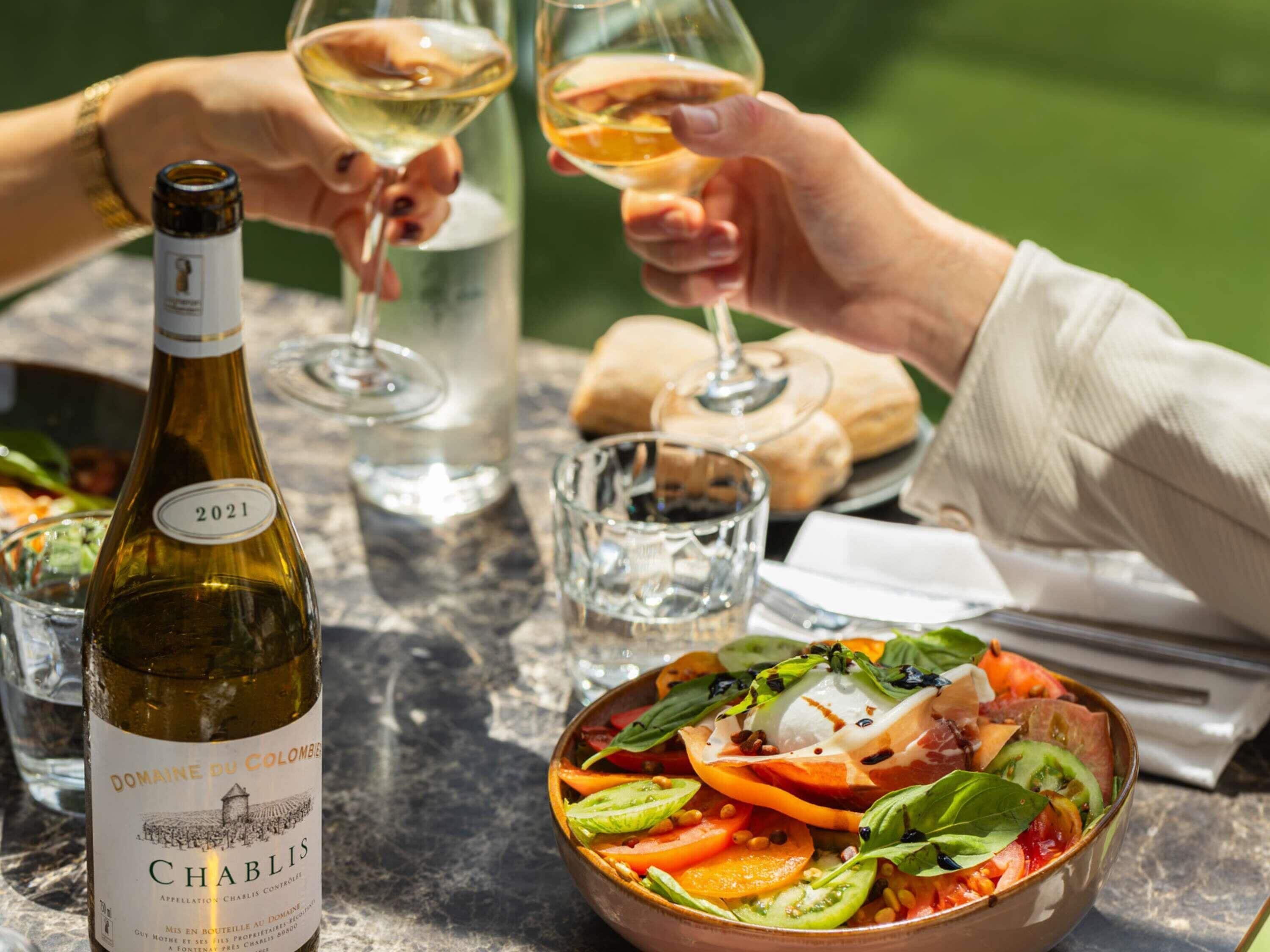 Una bottiglia di vino Chablis Domaine du Colombier 2021, una ciotola di insalata caprese e due bicchieri di vino bianco su un tavolo.