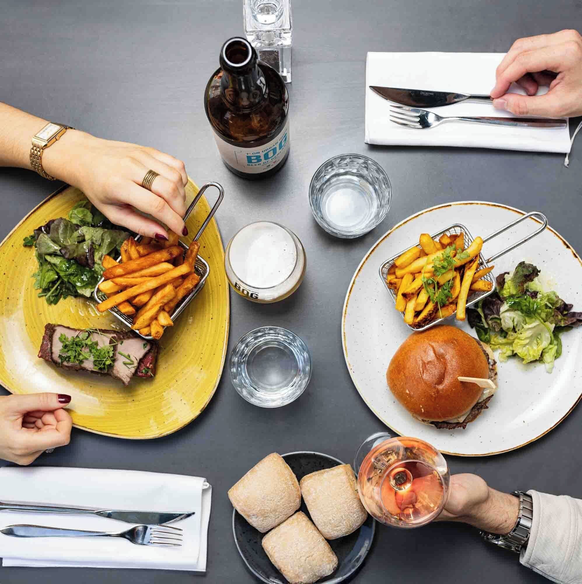 Una mesa de comedor con platos de comida, incluyendo bistec, papas fritas y una hamburguesa, junto con vasos de cerveza y agua.