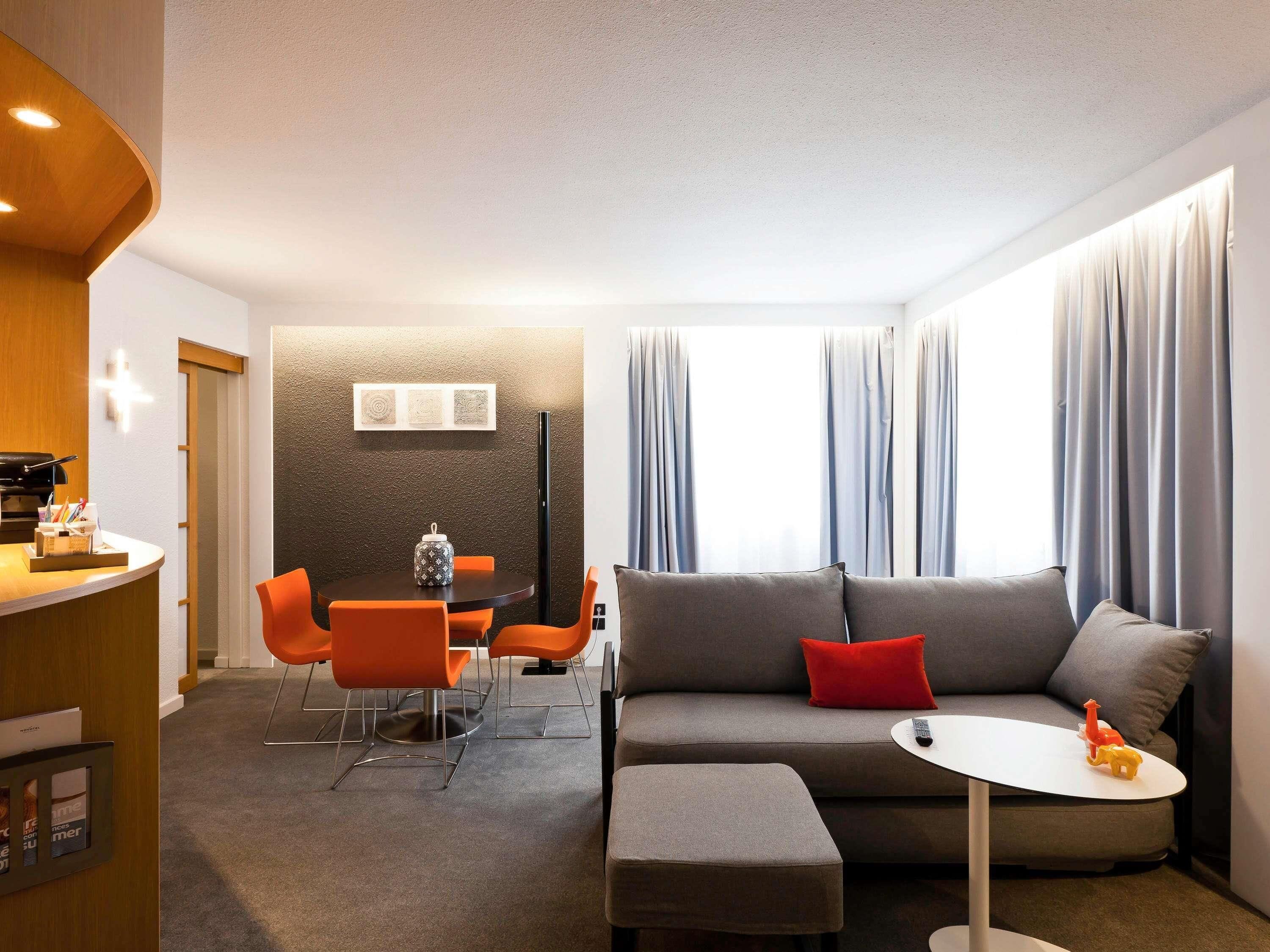 A modern living room with a sofa, ottoman, and a small table. There is a dining area with orange chairs and a table. The room has a large window with curtains.