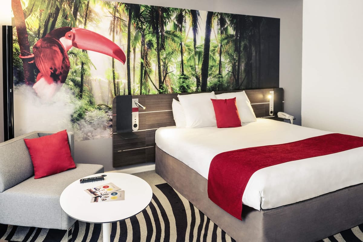 A modern hotel room with a large bed, a sofa, a small round table, and a wall mural of a forest scene.