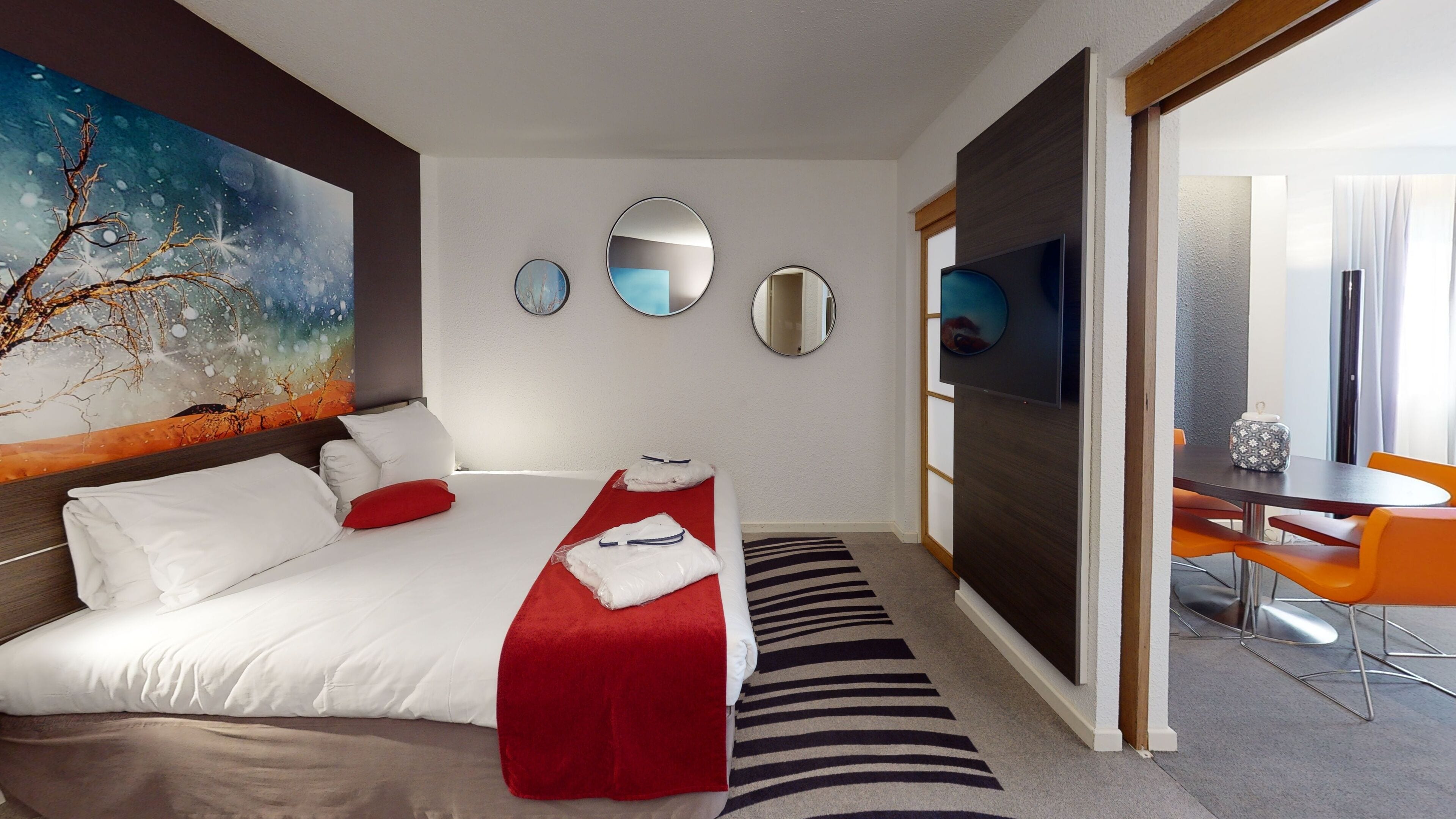 Una habitación de hotel con una cama grande, una colcha roja, un tapete a rayas y una zona de comedor con sillas anaranjadas.