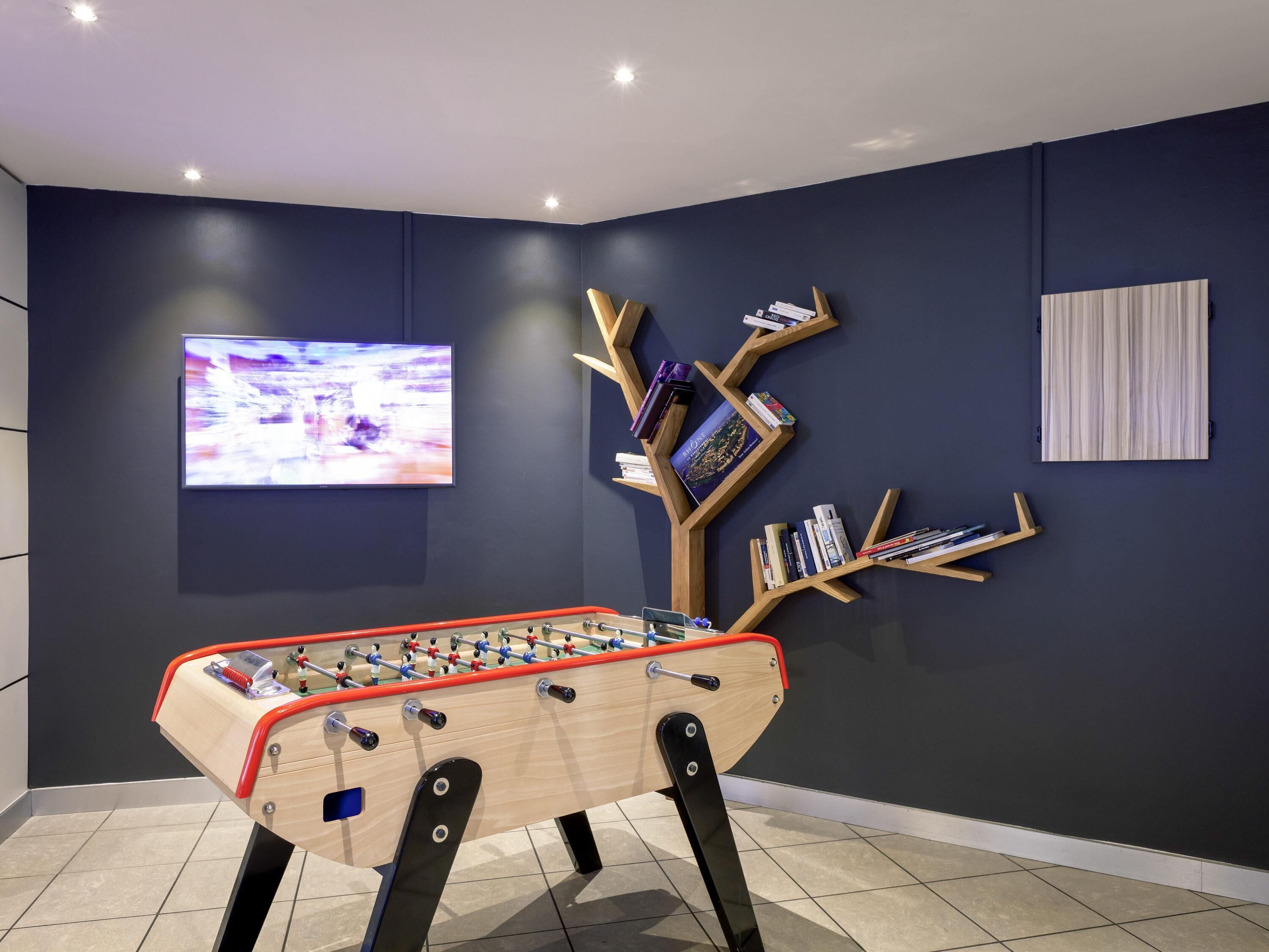A room with a foosball table, a wall-mounted TV, and a bookshelf with books and decorative items.
