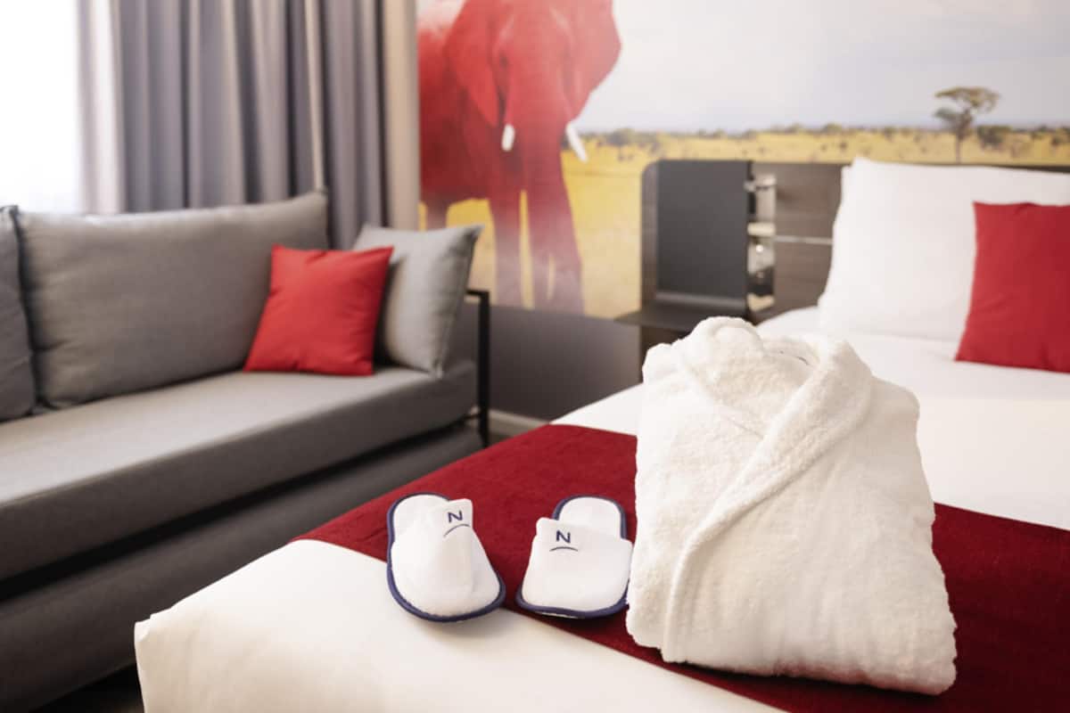 A hotel room with a bed, a sofa, a red throw blanket, and slippers.