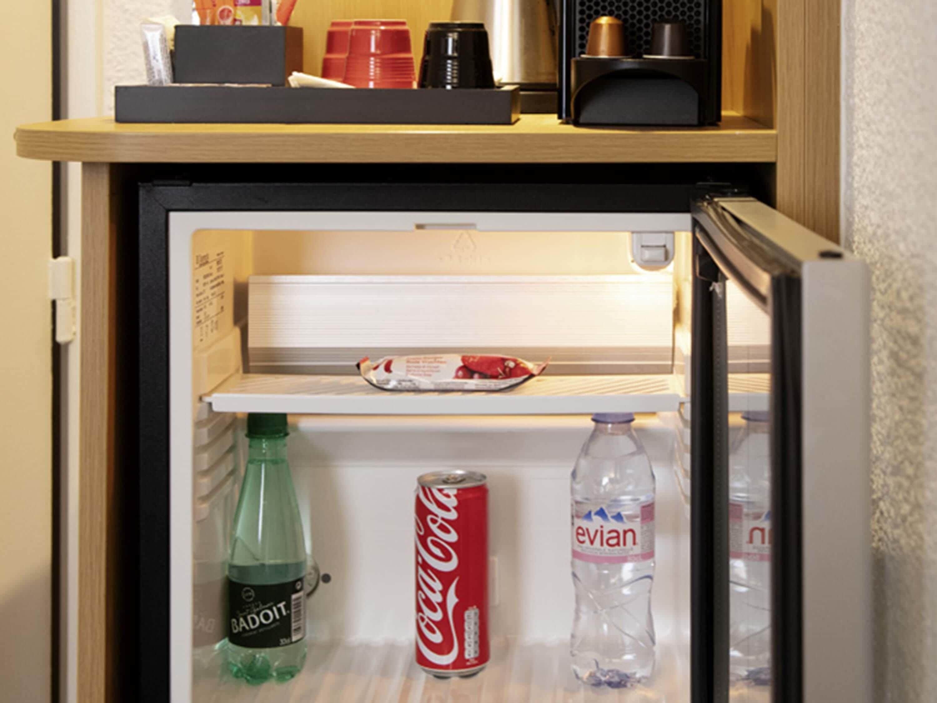 Un réfrigérateur avec une étagère contenant une bouteille de Badoit, une canette de Coca-Cola et une bouteille d’eau Evian.
