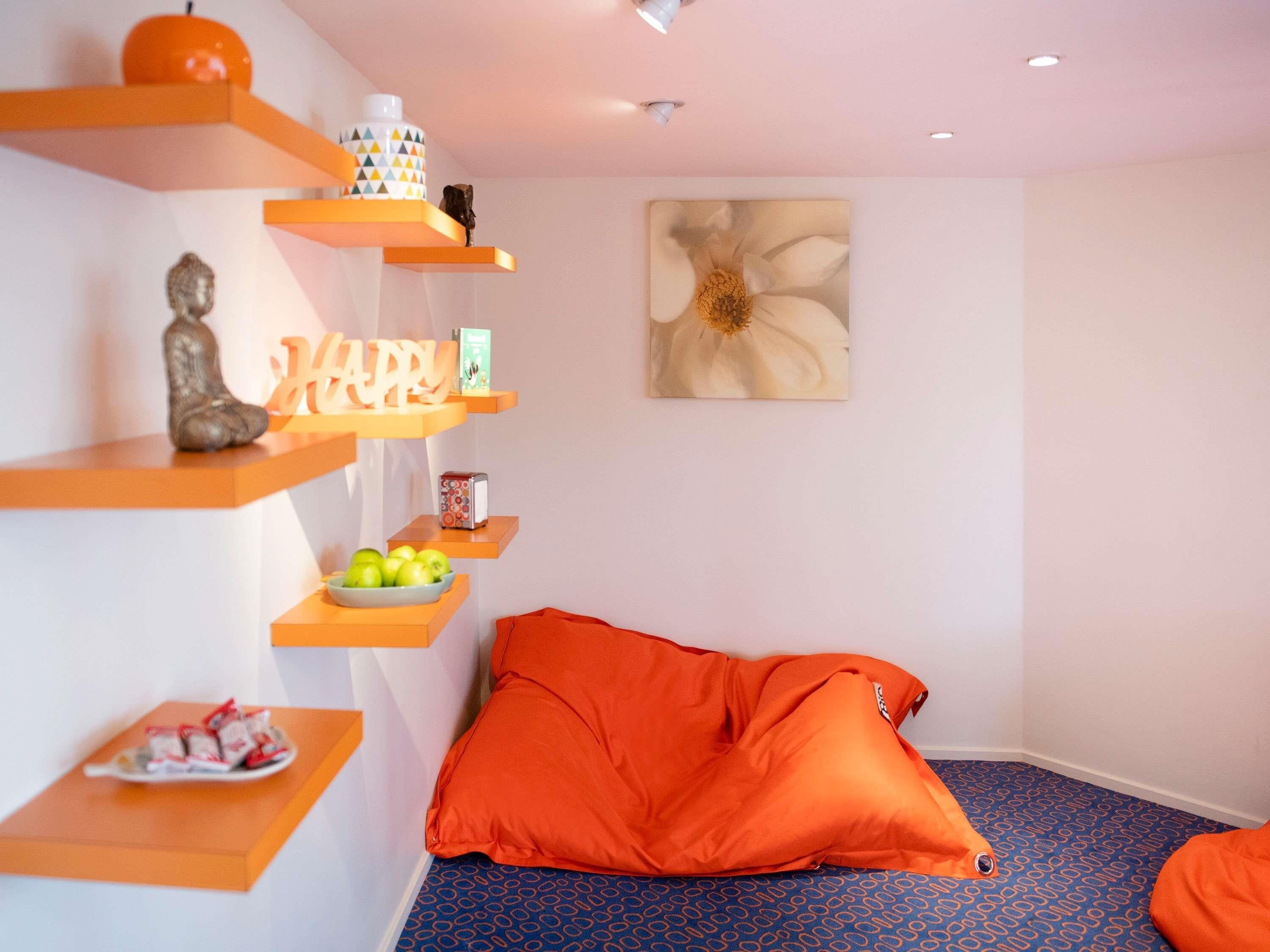 Een kamer met een blauw kamerbreed tapijt, oranje kussens, een Boeddhabeeld, een schaal appels, een boek en een bloemschilderij aan de muur.
