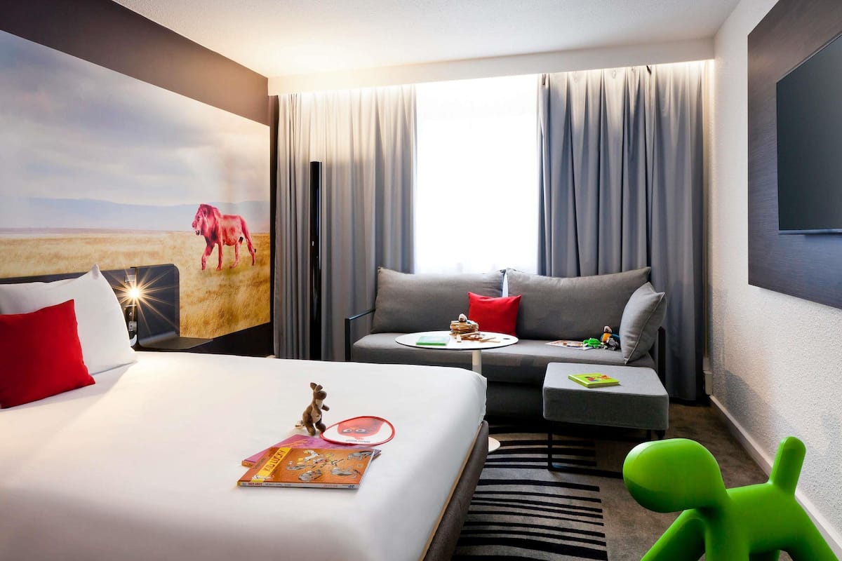 Habitación de hotel con cama, sofá, una mesita, una silla verde, un cuadro en la pared y una ventana con cortinas.