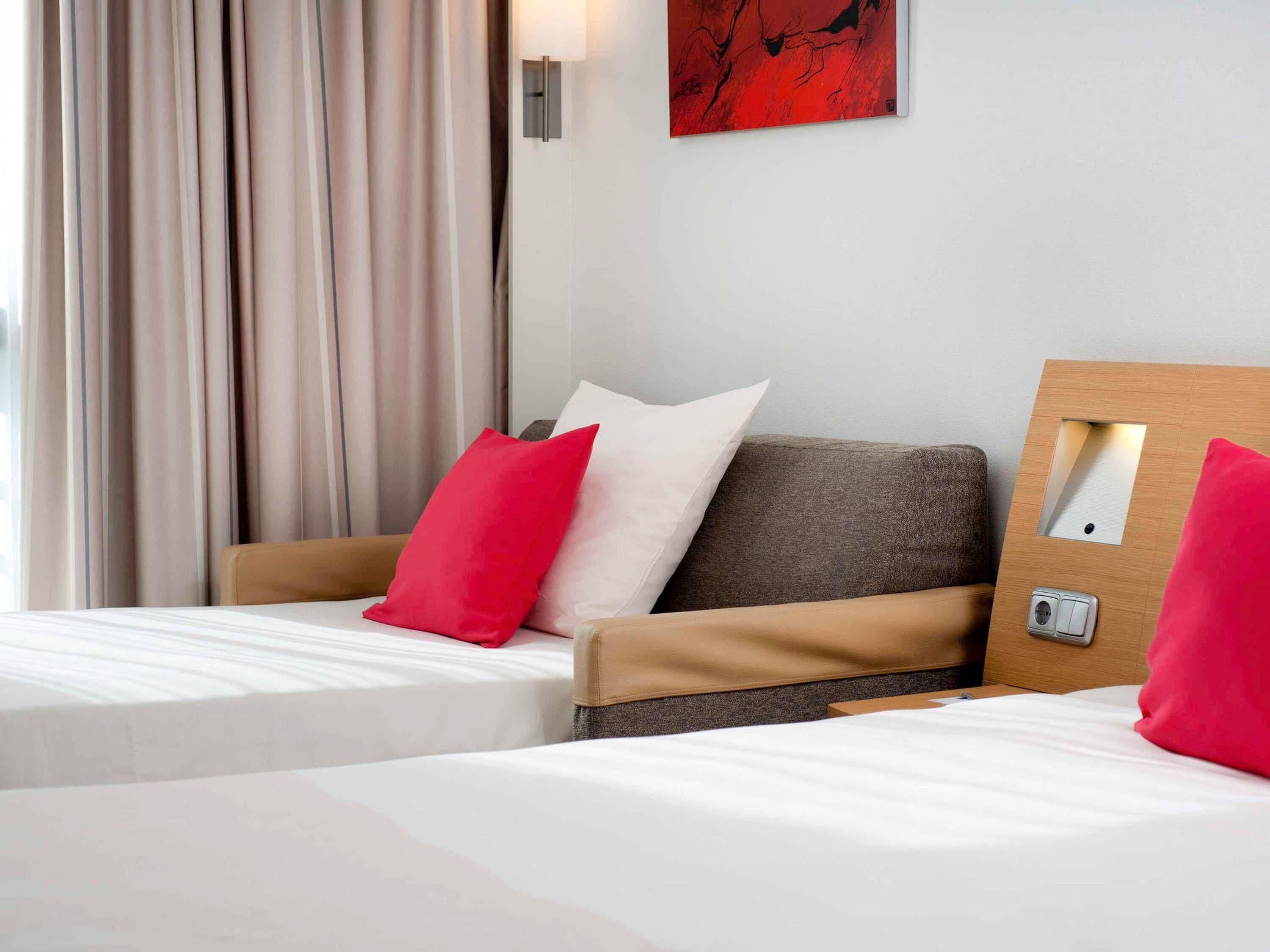 Hotelkamer met twee bedden, beige en bruine hoofdeinden, wit beddengoed, rode en witte kussens, een abstract schilderij in rood en wit aan de muur, en een houten hoofdeinde met spiegel en lichtschakelaar.