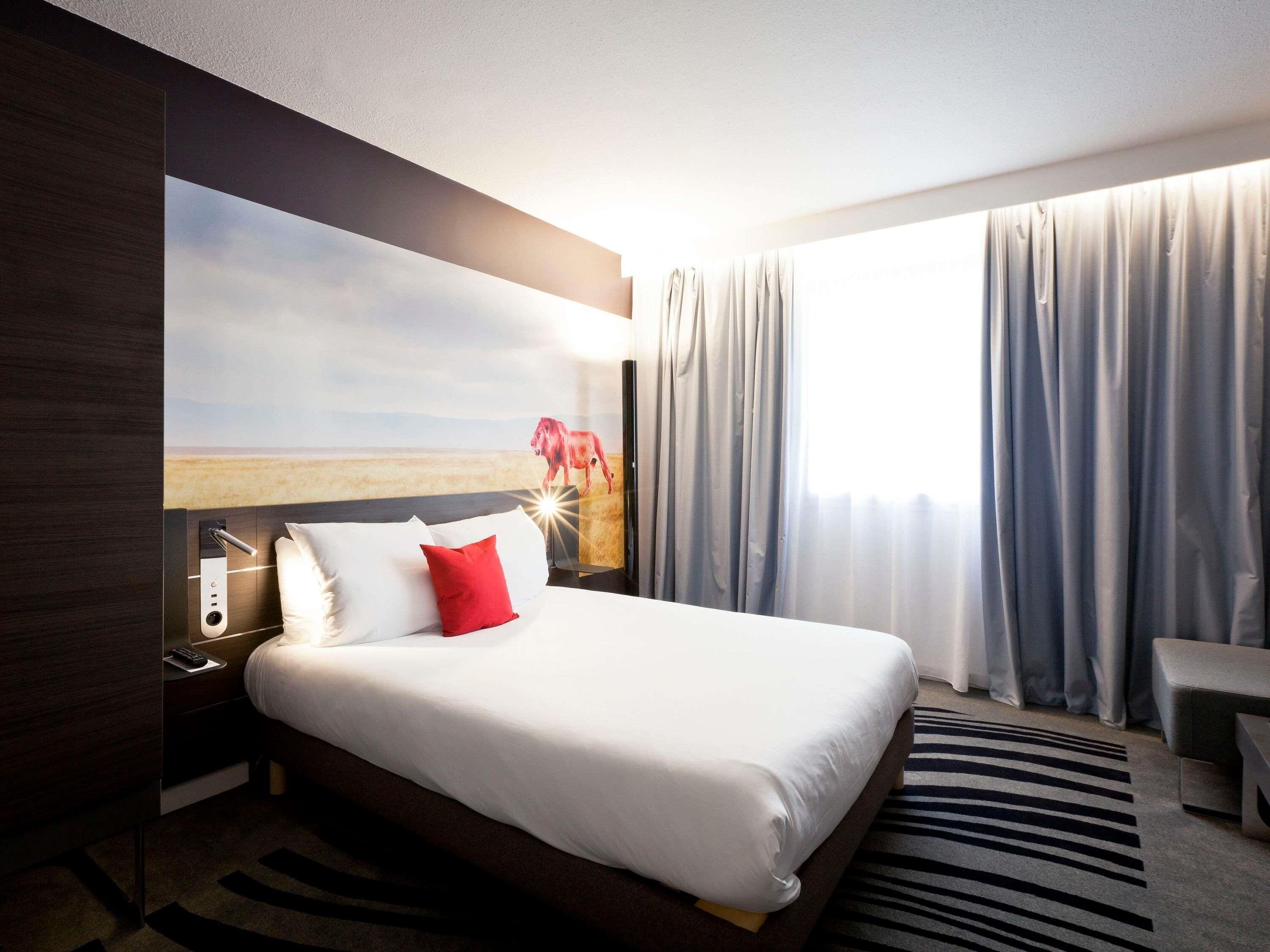 Ein Hotelzimmer mit Bett, Nachttisch, Lampe, einem Landschaftsbild und Blick ins Freie durch ein Fenster mit Vorhängen.