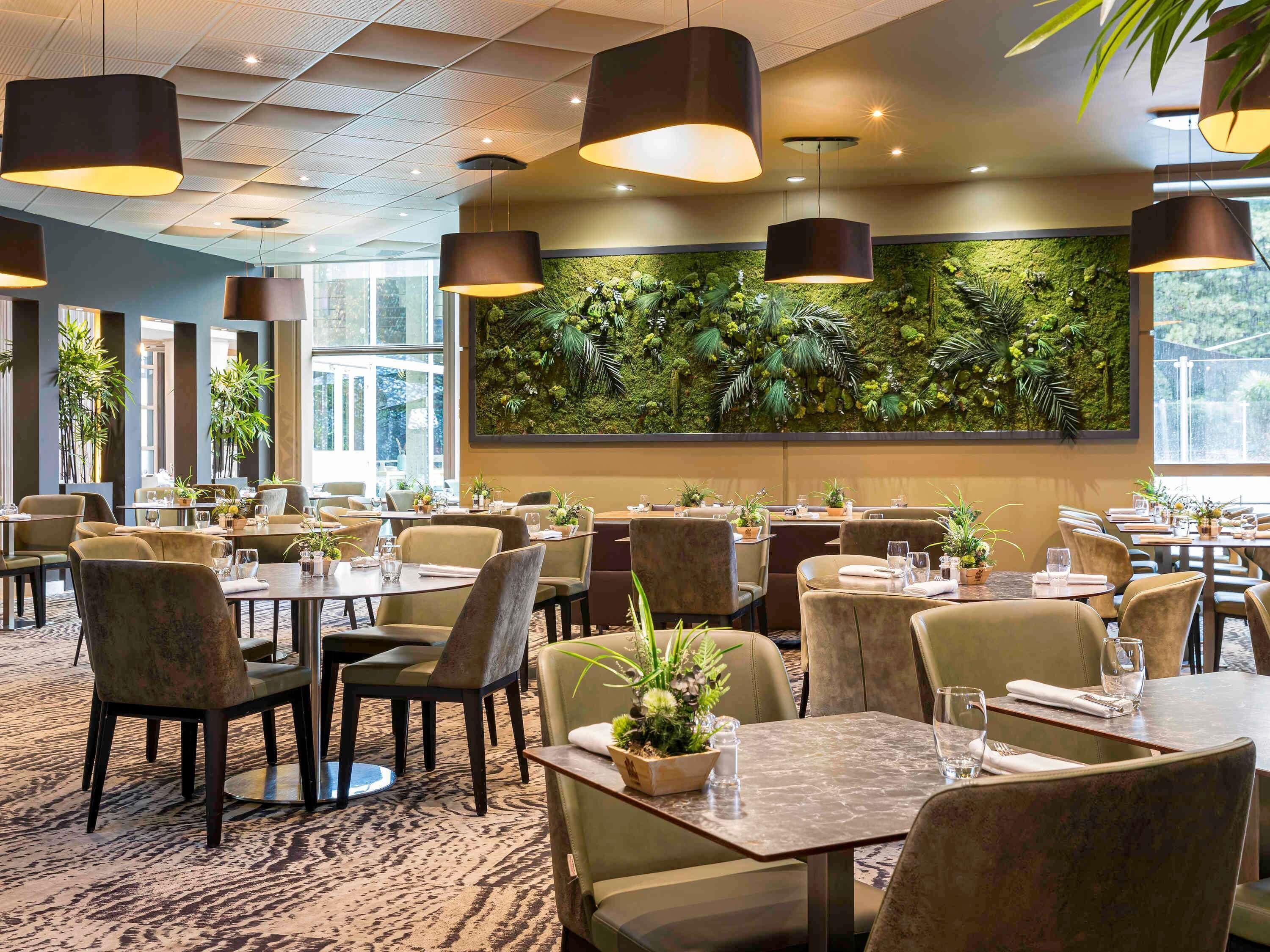 Un restaurante con mesas redondas, sillas, lámparas colgantes y una pared verde con plantas.