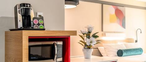 A modern hotel room with a kitchenette area featuring a Keurig coffee maker, a microwave, and a coffee pod holder. There is a bed with white bedding and a decorative pillow, a small potted plant, and a painting on the wall.