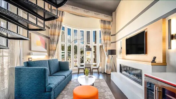 A modern living room with a blue sofa, a white coffee table, and an orange ottoman. There is a large window with curtains, a flat-screen TV mounted on the wall, and a fireplace. - Century Park Hotel (Los Angeles)