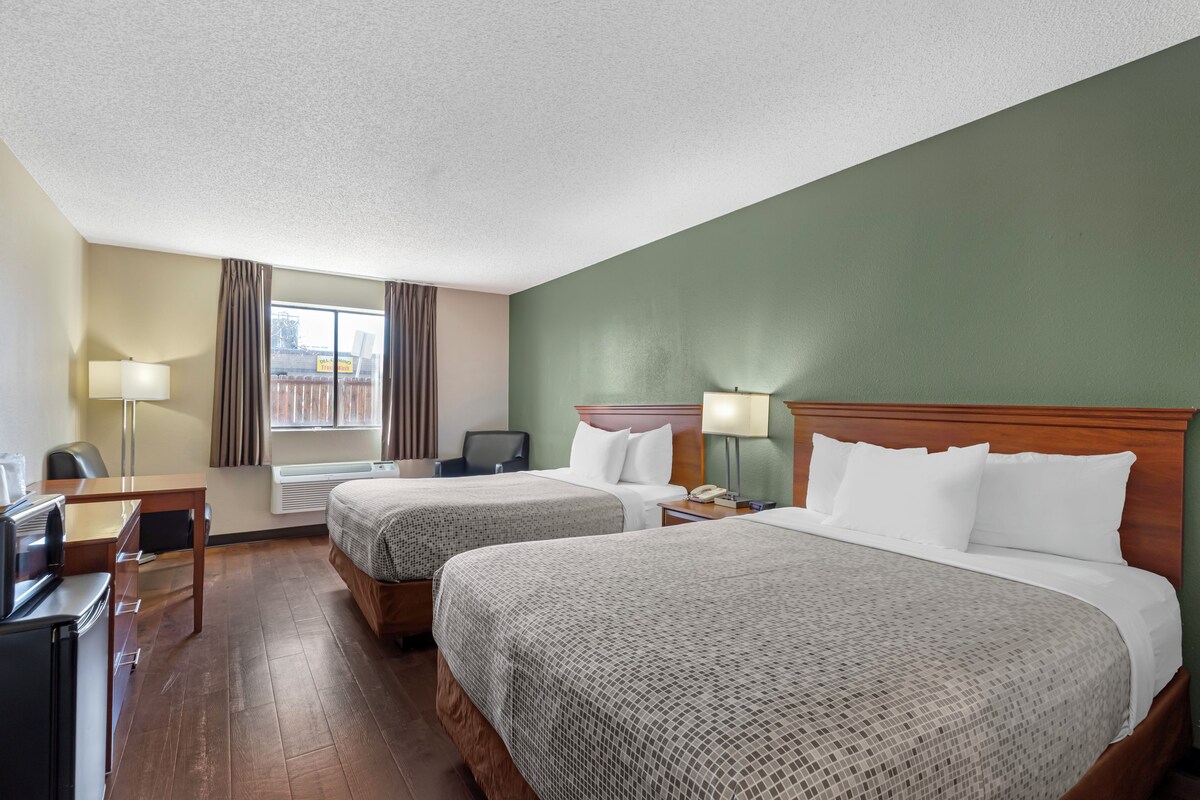 Quarto de hotel com duas camas, uma escrivaninha, uma cadeira, televisão e janela com cortinas.