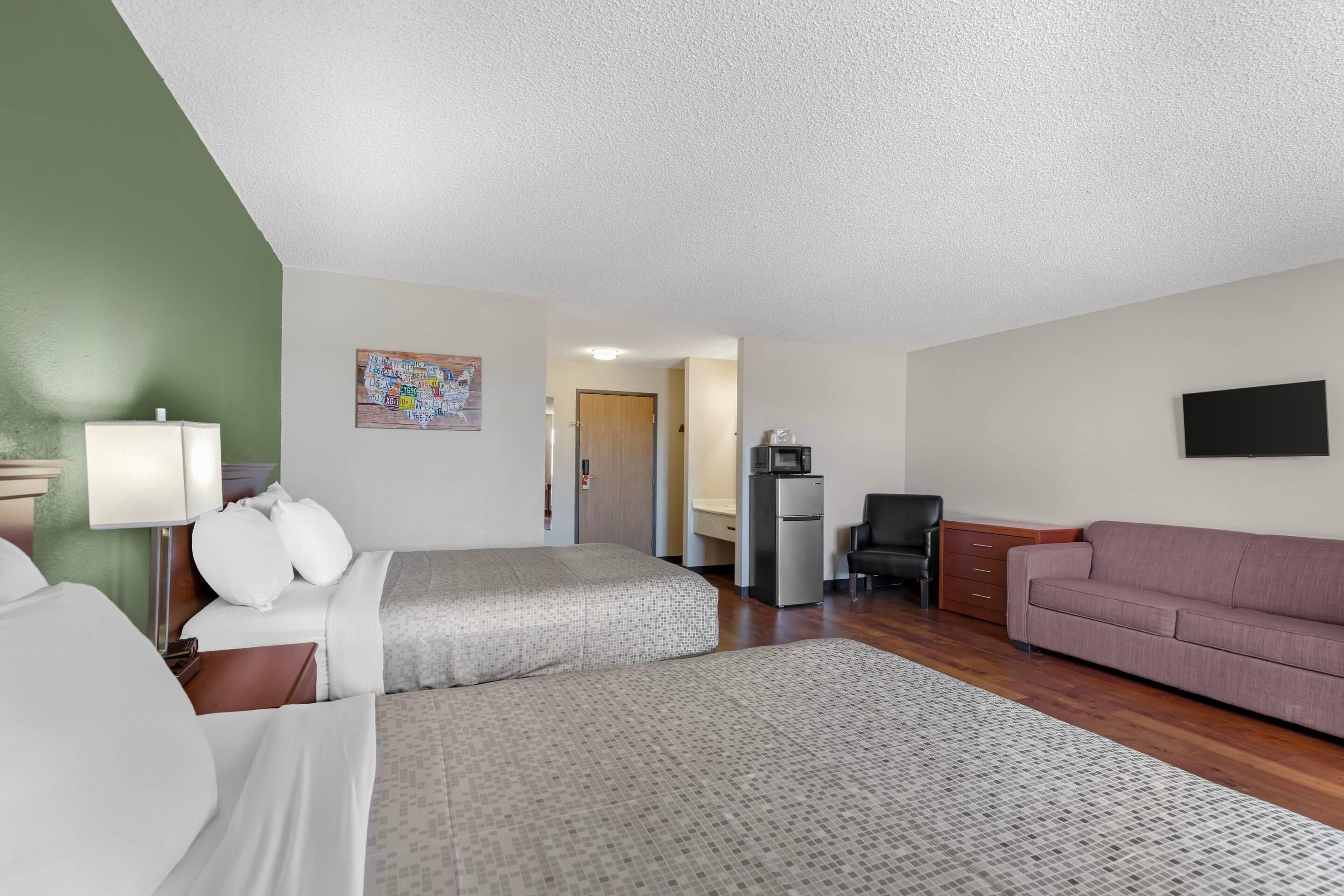 A hotel room with two beds, a sofa, a desk with a chair, a refrigerator, and a television on the wall.
