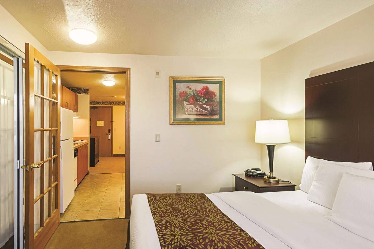A hotel room with a bed, a nightstand, a lamp, a painting, and a view of the hallway and kitchen.
