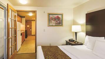 Quarto de hotel com uma cama, mesa de cabeceira, luminária, pintura e vista para o corredor e cozinha.