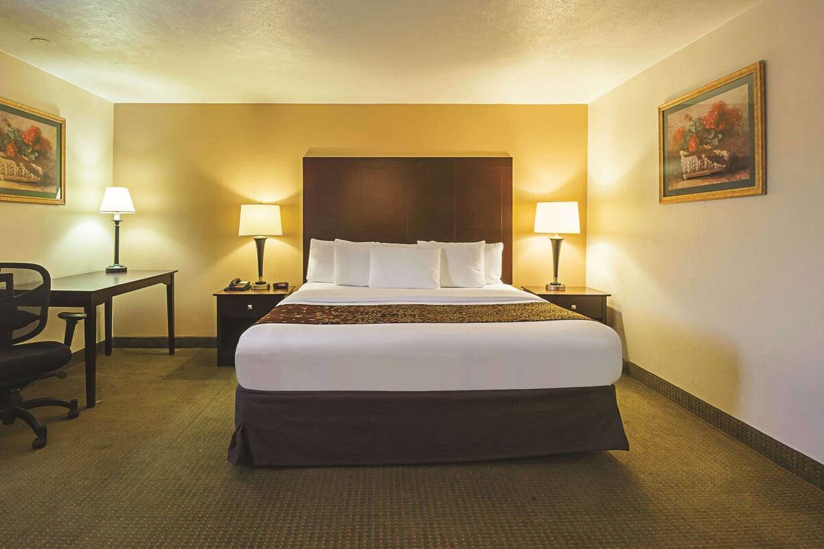 A hotel room with a large bed, two bedside tables with lamps, a desk with a chair, and two framed pictures on the walls.