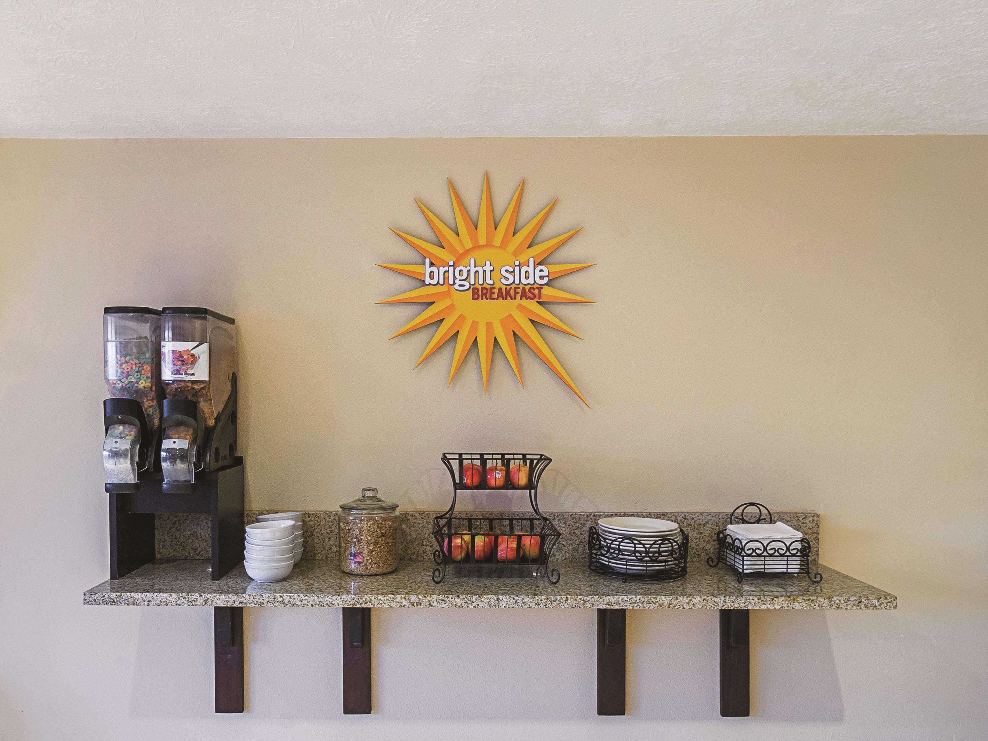 A breakfast bar with a coffee machine, cereal dispenser, fruit basket, and stacked plates.
