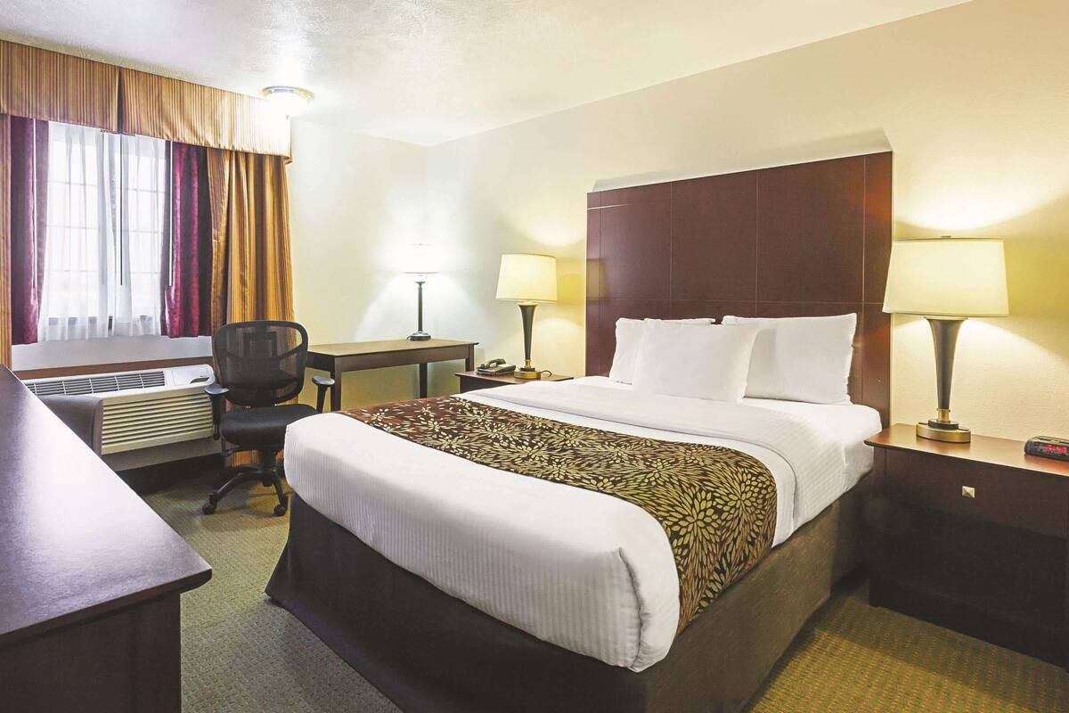 A hotel room with a bed, two bedside lamps, a desk with a chair, and an air conditioning unit.