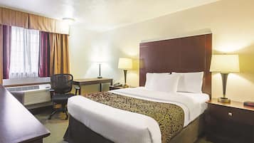 A hotel room with a bed, two bedside lamps, a desk with a chair, and an air conditioning unit.