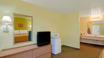 A hotel room with a bed, a television, a microwave, a refrigerator, a vanity with a mirror, and a lamp.