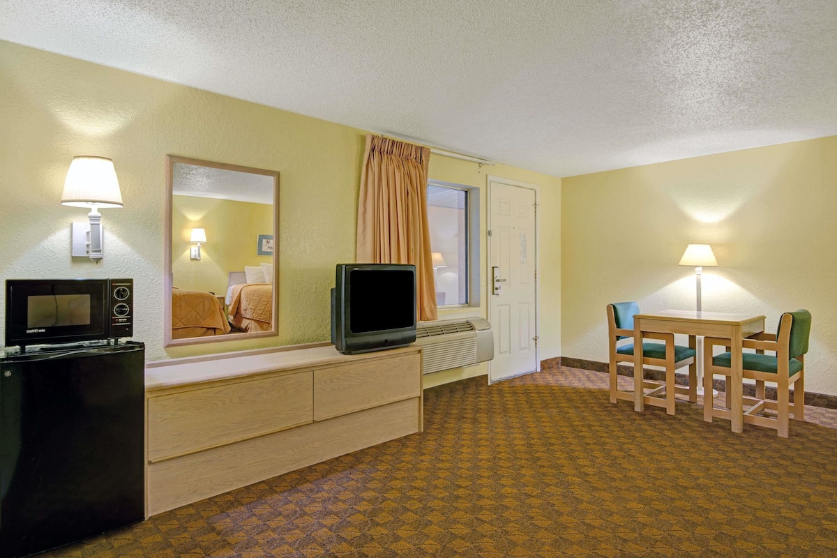 A hotel room with a bed, a desk with a computer, a television, a mirror, a window with curtains, and a dining area with chairs and a table.
