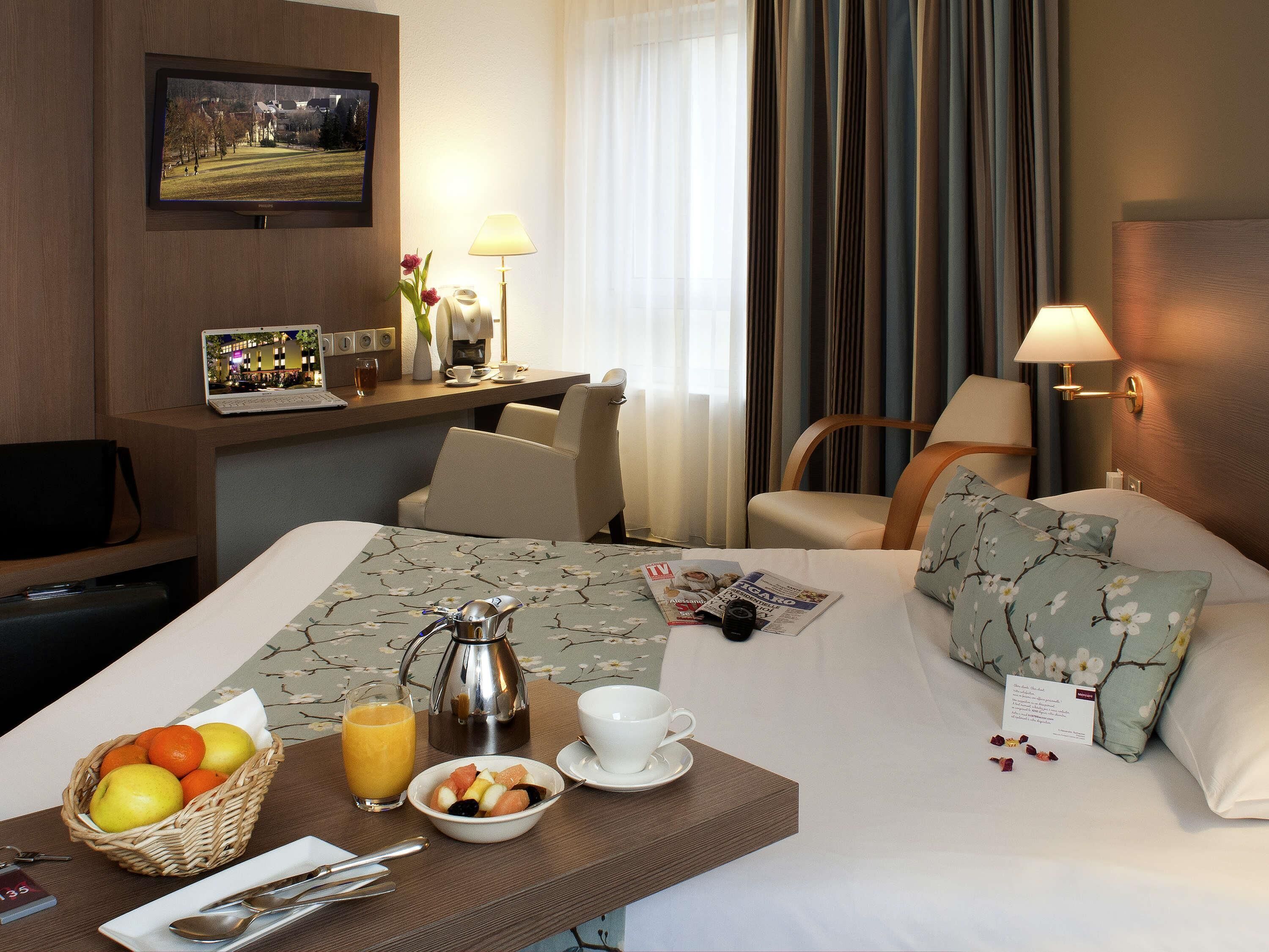 A hotel room with a bed, a desk with a laptop, a coffee maker, and breakfast items.