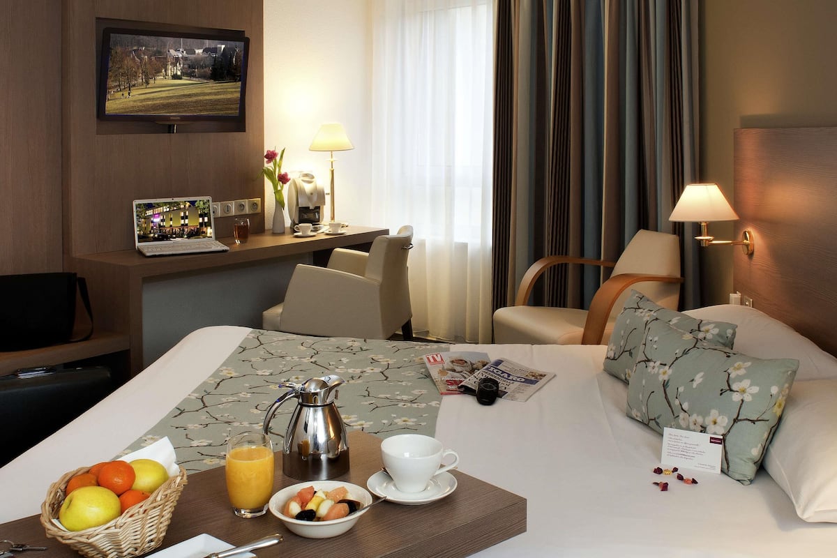 A hotel room with a bed, a desk with a laptop, a coffee maker, and breakfast items.