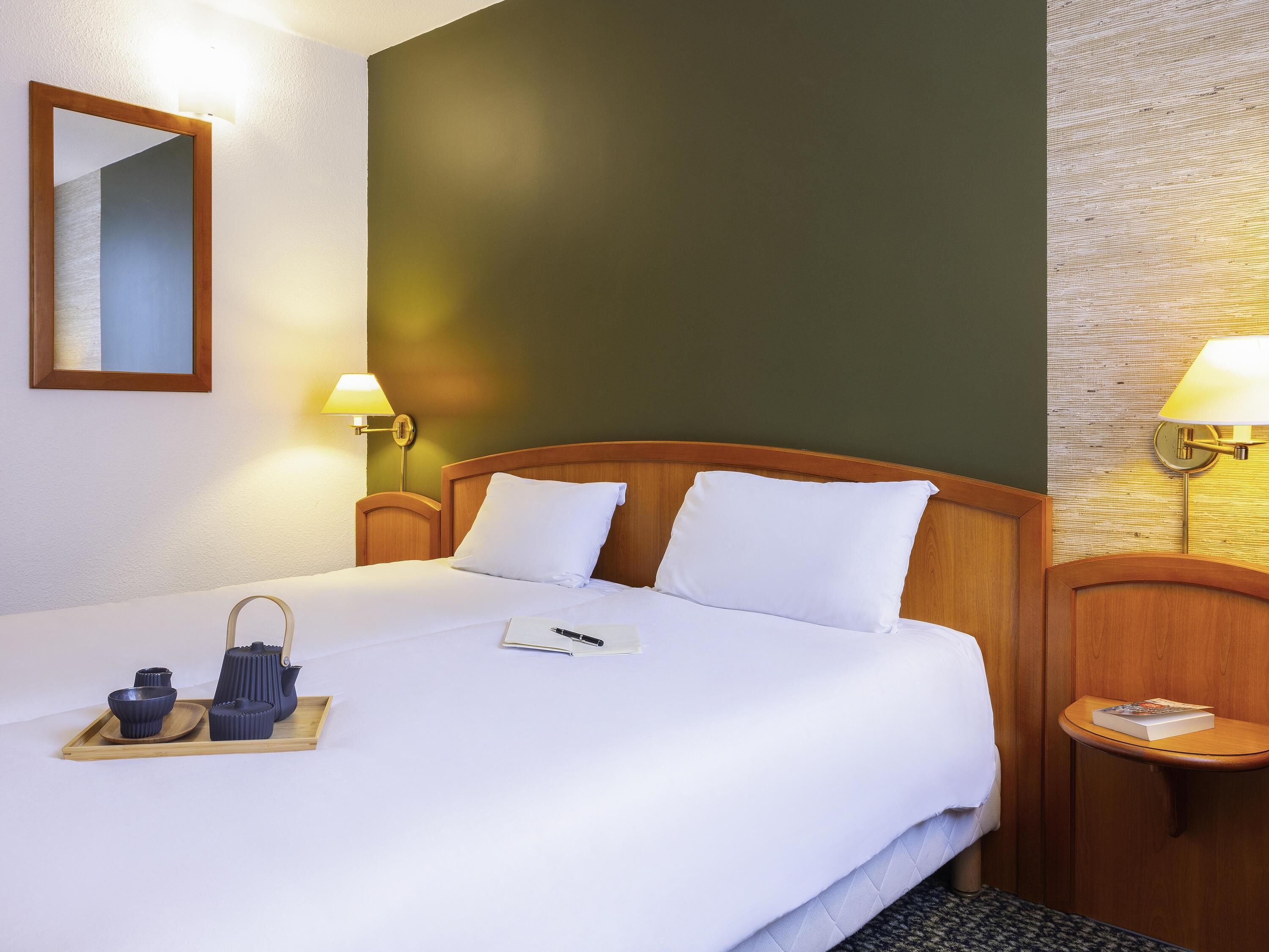 A hotel room with a bed, bedside tables, a tray with cups and a kettle, and a wall-mounted lamp.