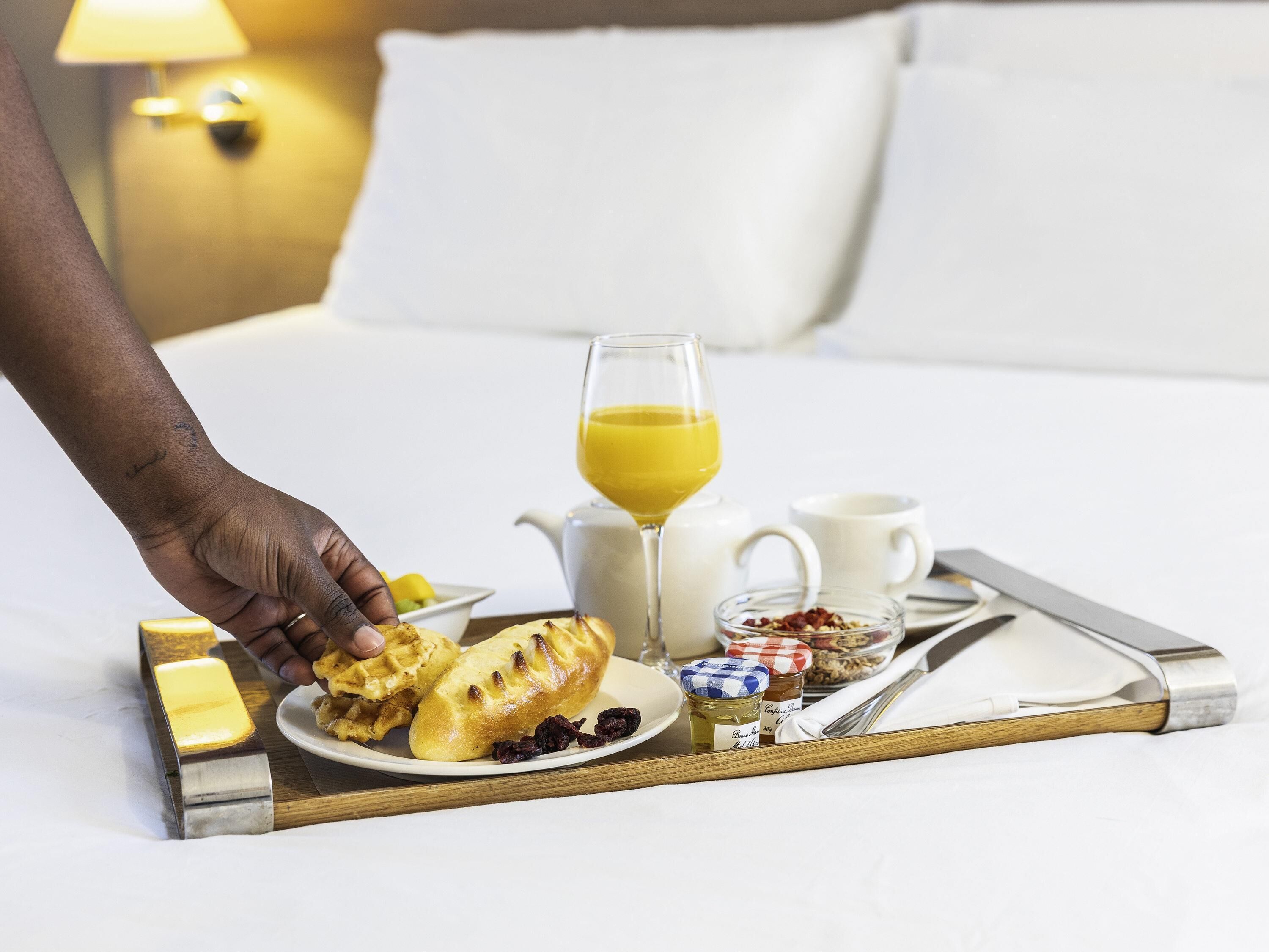 A breakfast tray with waffles, a glass of orange juice, a jug, a cup, and a small jar of jam on a bed.