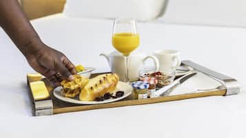 A breakfast tray with waffles, a glass of orange juice, a jug, a cup, and a small jar of jam on a bed.