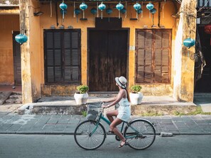 Une femme fait du vélo dans une rue, devant un immeuble orné de volets en bois et de plantes en pot.