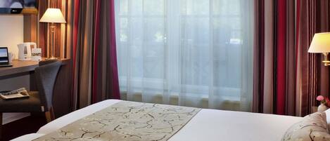 A hotel room with a bed, a desk with a laptop, a television, and a window with curtains.
