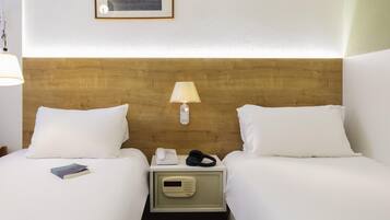 Habitación de hotel con dos camas, una mesita de noche con lámpara, teléfono y reloj digital.