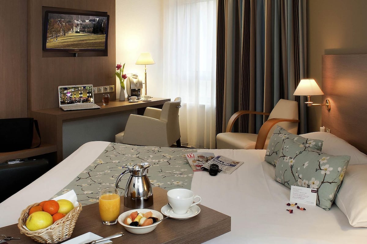 A hotel room with a bed, bedside table, and a desk with a laptop, lamp, and flowers.