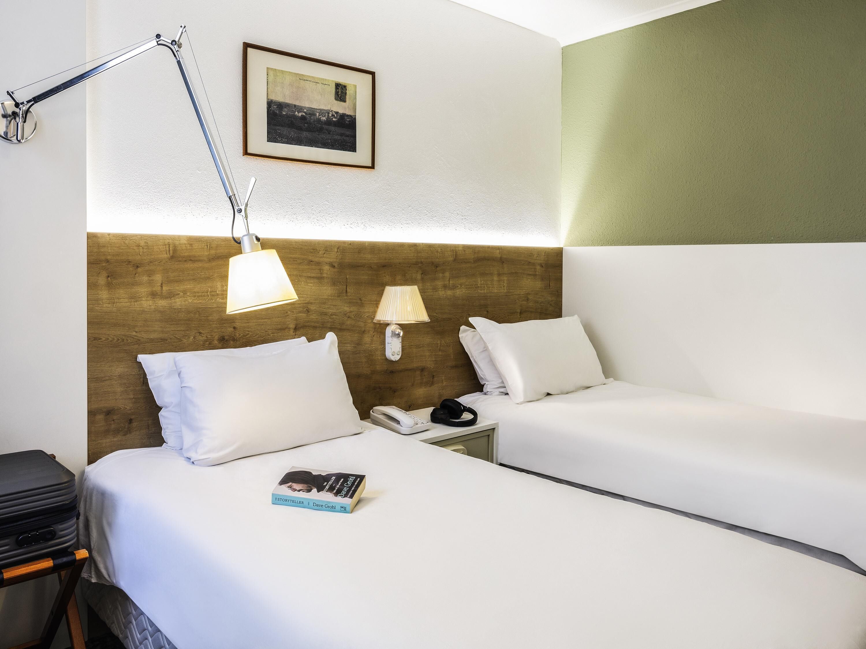 Ein Hotelzimmer mit zwei Betten, einem hölzernen Kopfteil, Nachttischlampen, einem gerahmten Bild an der Wand, einem Telefon am Nachttisch und einem Buch auf dem Bett links.