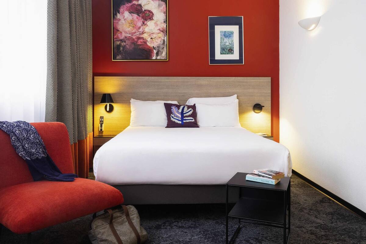 A hotel room with a bed, a chair, a nightstand, a lamp, and two wall-mounted light fixtures.