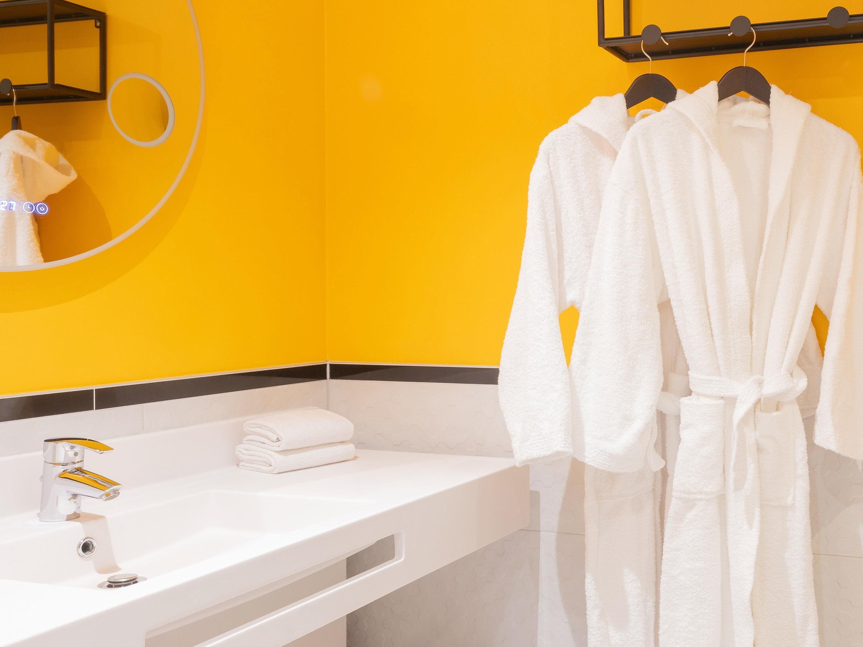 A bathroom with a yellow wall, a white sink, and two white robes hanging on a rack.