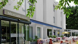 A Mercure Hotel exterior with a glass entrance, outdoor seating area with tables and chairs, and a signboard. - Mercure Forbach (Forbach)