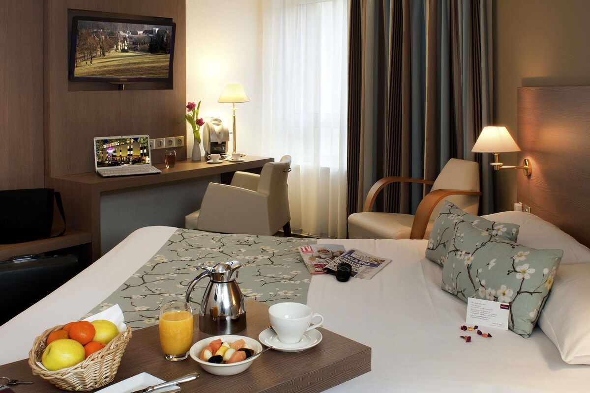 A hotel room with a bed, a desk with a laptop, a coffee maker, and breakfast items.