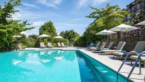 A swimming pool with lounge chairs and umbrellas, surrounded by trees and a stone building. - Hôtel Ermitage (Neuvecelle)