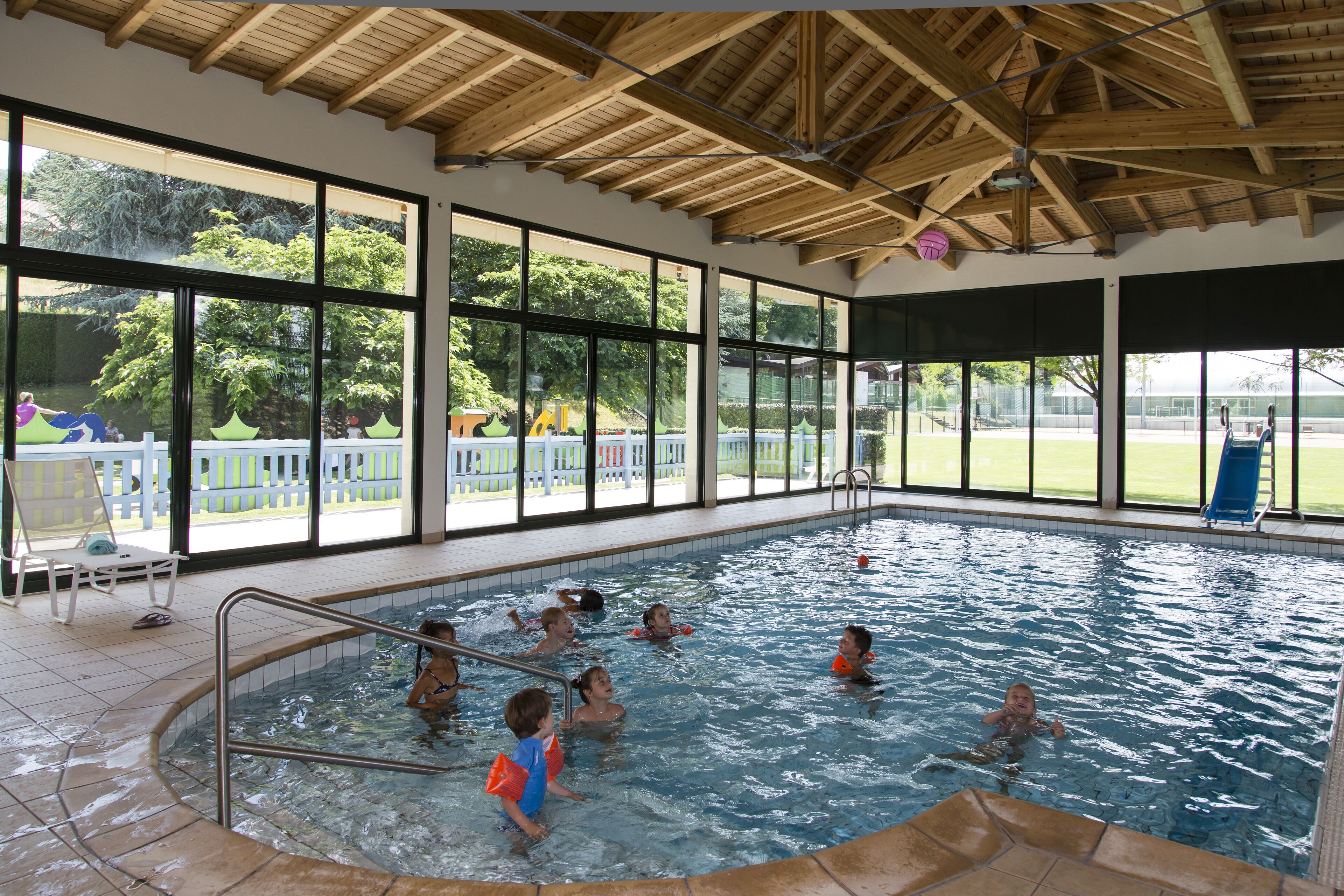 Una piscina interior con vistas despejadas al exterior, techo de madera y zona junto a la piscina con sillas.