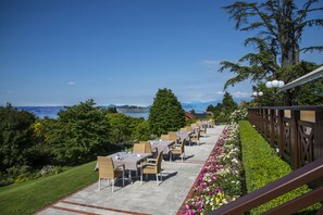 An outdoor dining area with tables and chairs, overlooking a scenic landscape with trees and a view of the sea.
